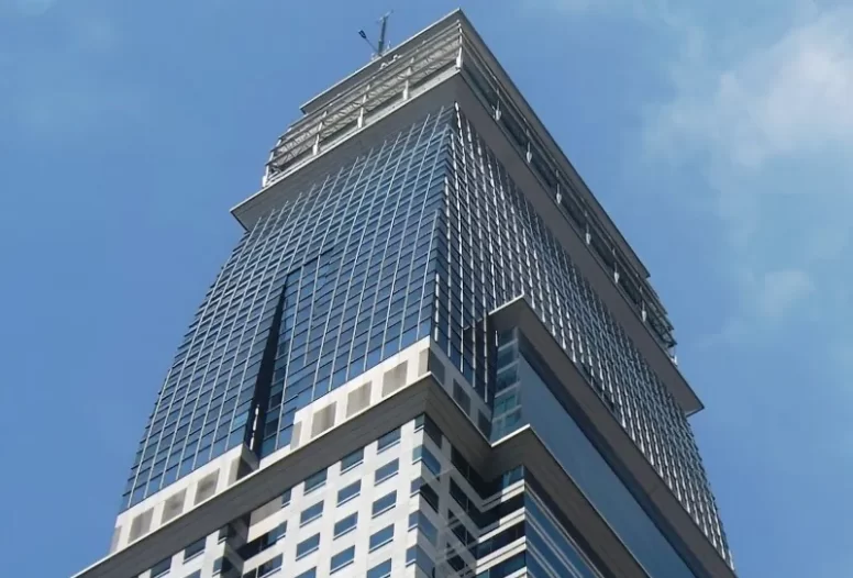 A ground level, upwards view of the top of the white, dark blue and gray steel and stone facade of Capital Tower, a high-rise building in Singapore.