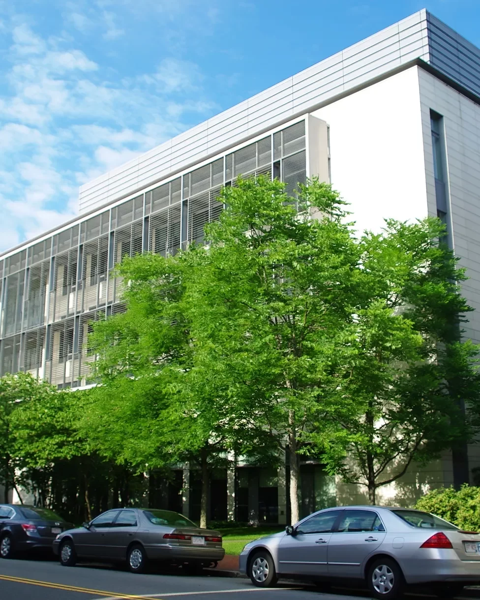 The dark gray, metal shading system set against the light gray facade of the Goel Quantum Science & Engineering Building surrounded by the green trees at Harvard University.