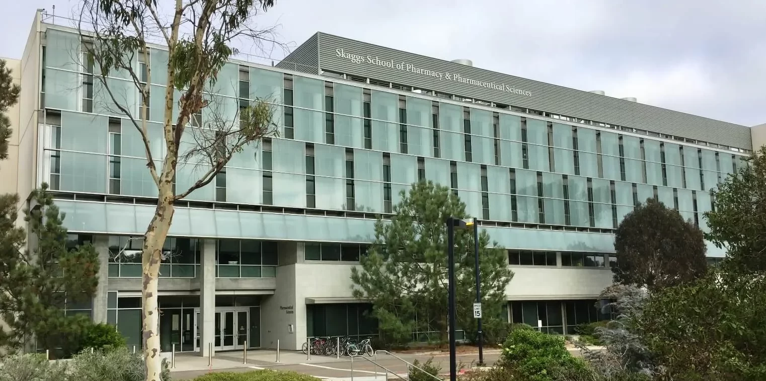 The external glass facade shades on the Skaggs School of Pharmacy at UC San Diego against a cloudy blue sky.