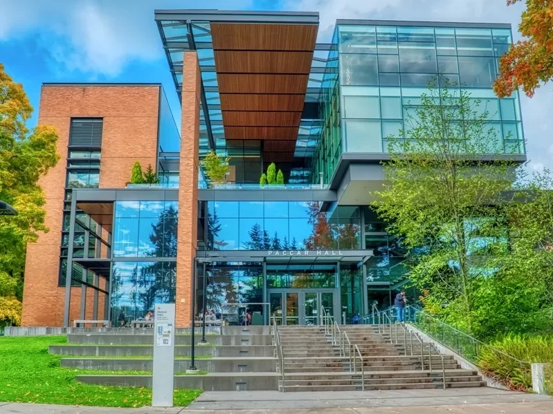 The front of PACCAR Hall at the University of Washington, showing the cantilevered gray steel, green glass, and red brick facade.