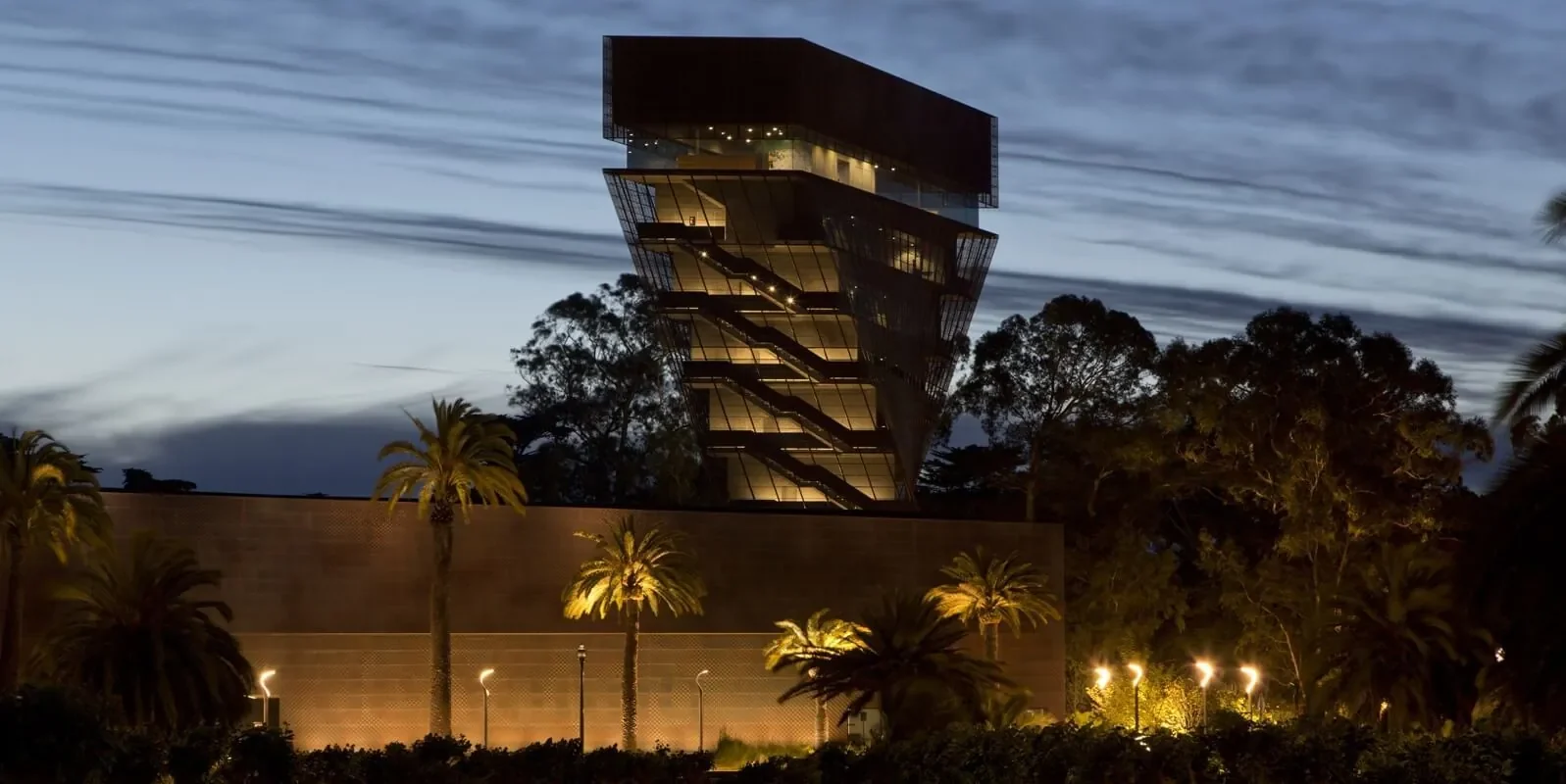 Sunset view of the spotlight-lit twisted metal tower of the de Young Museum set against a cloud-streaked sky.