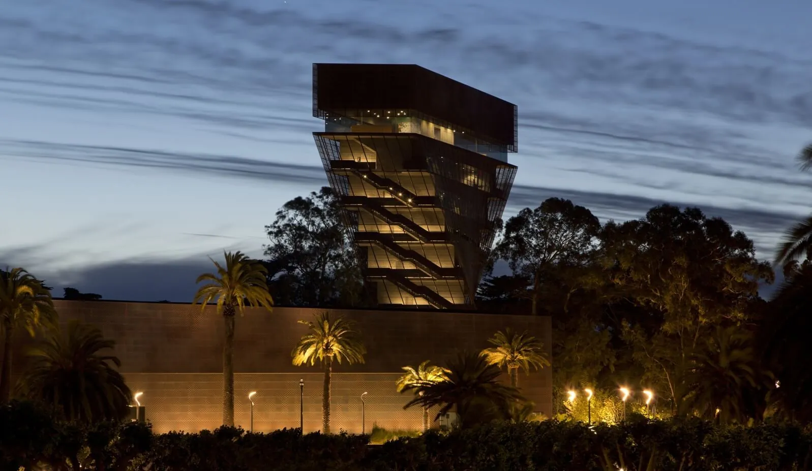 Sunset view of the spotlight-lit twisted metal tower of the de Young Museum set against a cloud-streaked sky.