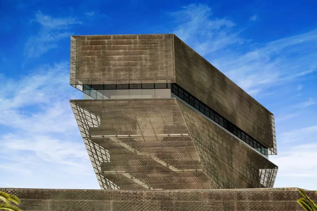 The punctured brown metal facade of the tower of the de Young Museum against a cloudy blue San Francisco sky.