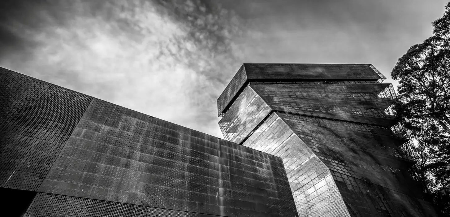 Black and white image looking up at the skewed tower of the de Young Museum from ground level.