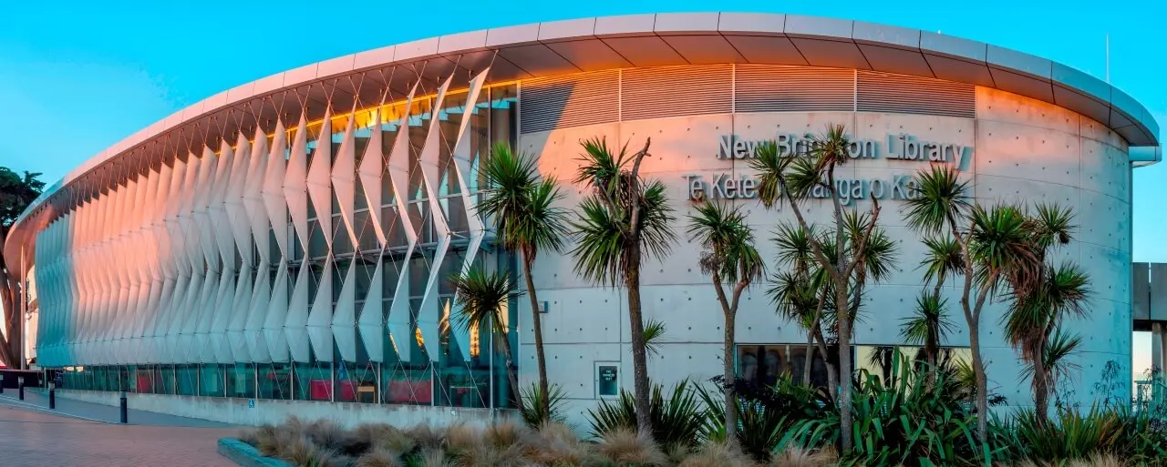The concrete & fabric sail facade of the New Brighton Library in Christchurch, NZ in front of a bright turquoise sky.