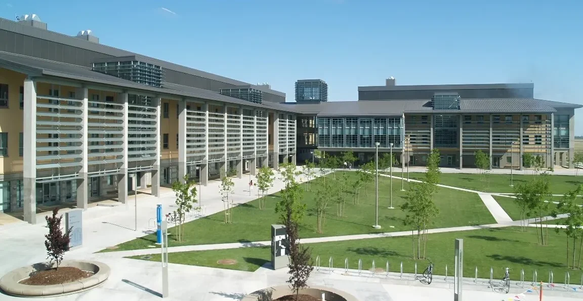 A panoramic view of the blue Science + Engineering building at UC Merced, in a sea of grass and young trees.