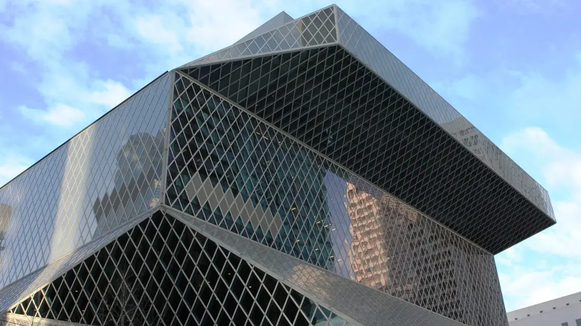 A close up of the black and silver geometric facade of the Seattle Central Library set against a cloudy blue sky.