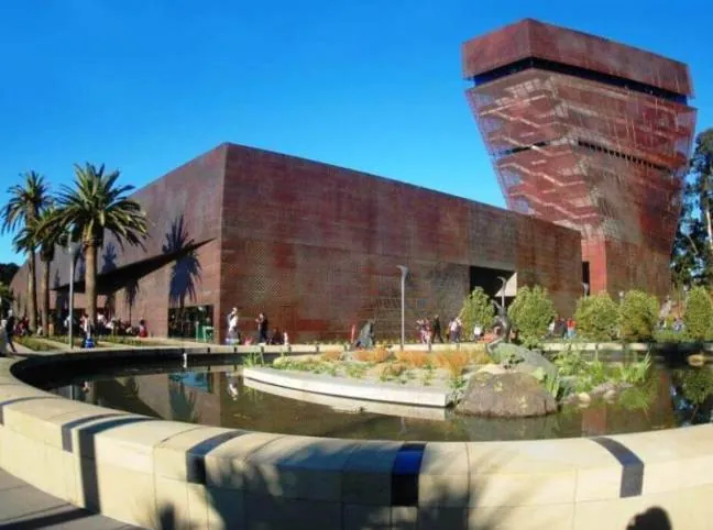 The punctured brown metal facade of the twisted tower and rectilinear building of the de Young Museum against a bright blue San Francisco sky.
