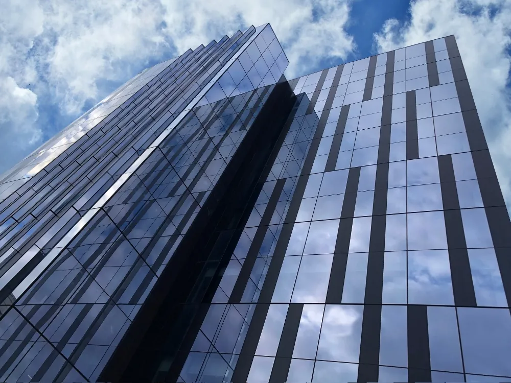 A ground level, upwards view of the blue glass & steel facade of Genentech's corporate office Building 40 against a cloudy California sky.